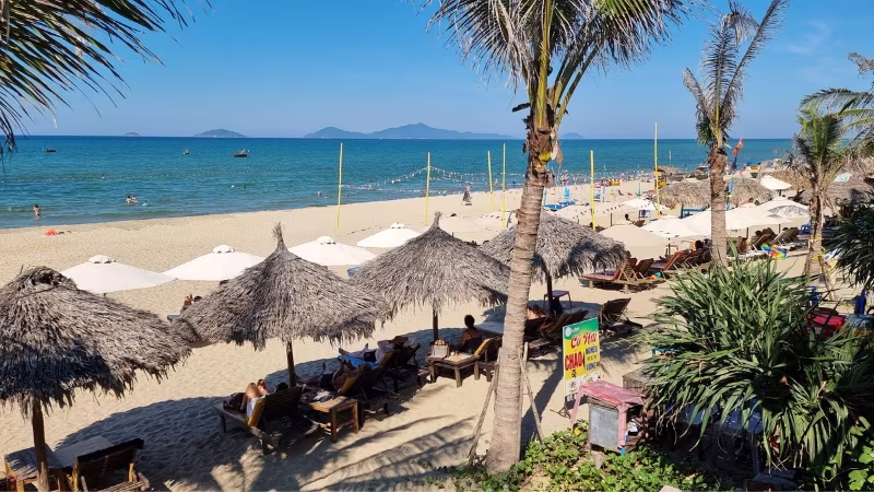 Sun loungers and thatched umbrellas lining An Bang Beach in Hoi An, with palm trees, white sand and calm blue sea under a clear sky