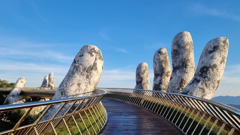 Golden Bridge in Da Nang supported by giant stone hands, curving through the Ba Na Hills under a clear blue sky

