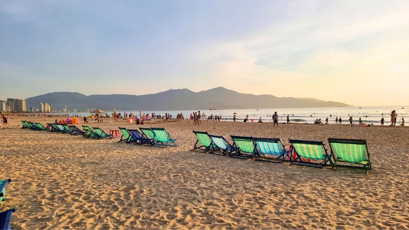 Long Stays in Vietnam Beachgoers relaxing on deck chairs at My Khe Beach in Da Nang, Vietnam during sunset with mountains in the background
