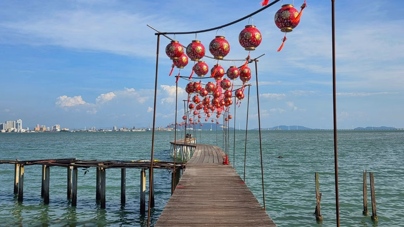 Wooden Lee Jetty decorated with red Chinese lanterns extending into the sea in George Town, Penang, with city skyline and blue sky in the background