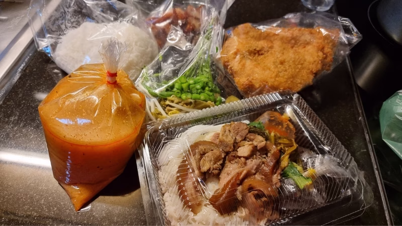 Takeaway Thai street food with rice, stewed pork, curry sauce, fried chicken cutlet and vegetables packed in plastic containers and bags