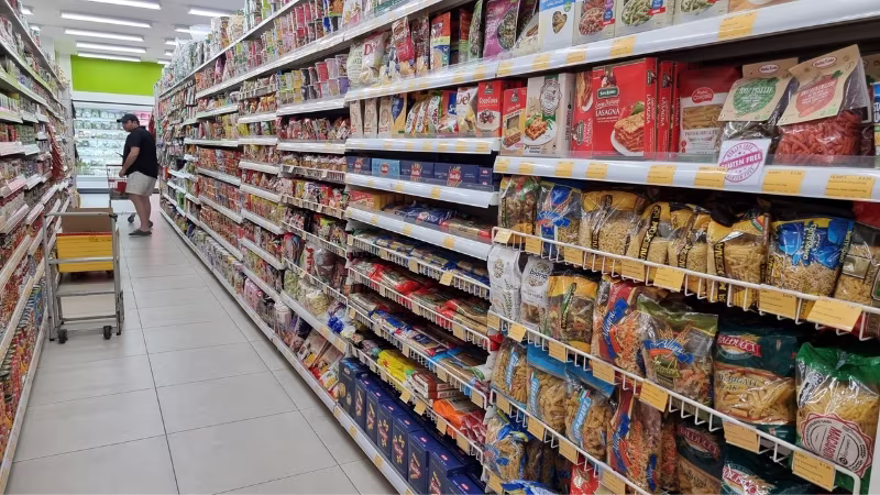 Super Duper grocery store aisle stocked with packaged pasta, sauces and dry goods, with a shopper browsing shelves and a yellow trolley nearby