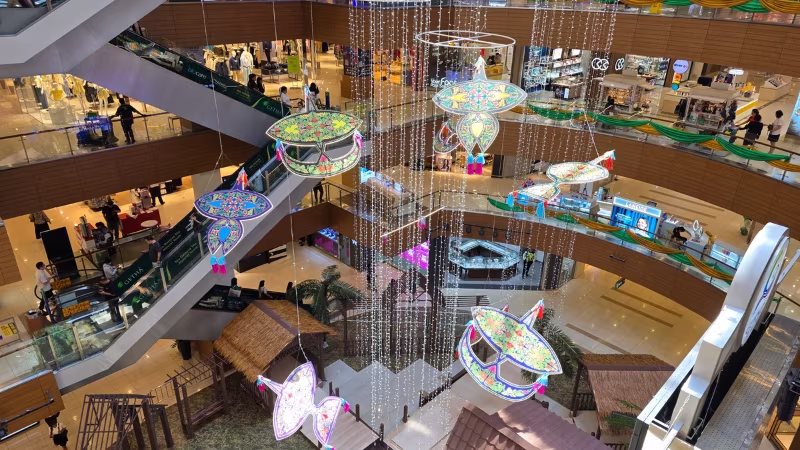 Gurney Plaza in Penang decorated with colourful hanging wau kites and festive lights for Hari Raya, surrounded by retail shops and escalators across multiple levels