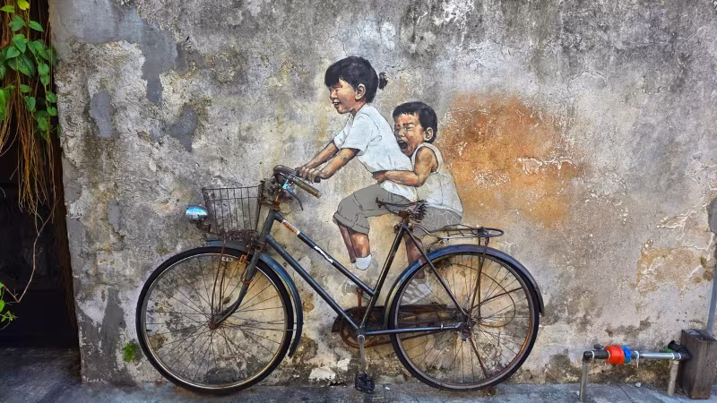 Street art mural of two children riding a real bicycle on Armenian Street, George Town, Penang, blending 3D object with painted artwork for optical illusion