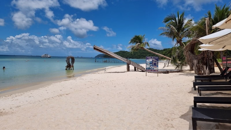 Cheapest Beach Destinations to Visit from Australia Wooden jetty and palm trees on white sand Sao Beach at Phu Quoc Island, Vietnam, with calm turquoise water and boat in the distance under blue sky